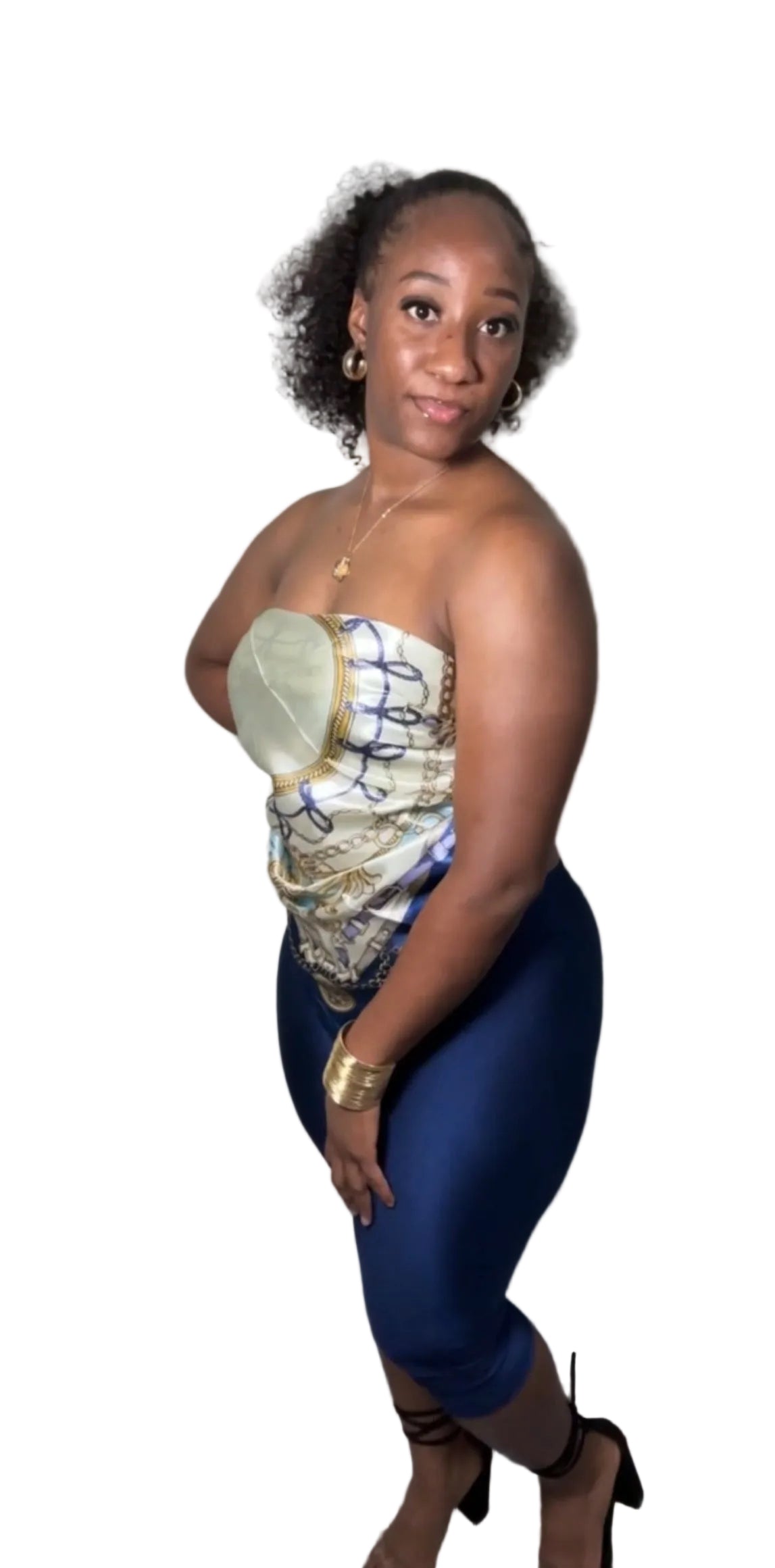 A woman posing indoors wearing a strapless top with a gold, white, and blue pattern, paired with dark blue leggings and black heels.